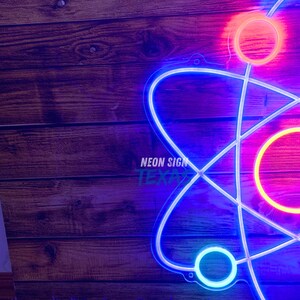 May include: A neon sign in the shape of an atom, with a blue and white outline. The sign has a bright pink and orange glow. The text "NEON SIGN TEXAS" is visible on the sign. The background is a wooden surface.