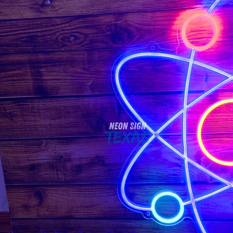 May include: A neon sign in the shape of an atom, with a blue and white outline. The sign has a bright pink and orange glow. The text "NEON SIGN TEXAS" is visible on the sign. The background is a wooden surface.