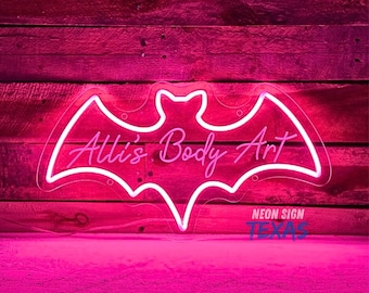 May include: Pink neon sign shaped like a bat with the text "Alli's Body Art" in white neon. The sign is on a pink wood background.