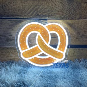 May include: A neon sign in the shape of a pretzel, with a white outline and a light-up orange and yellow interior. The sign is mounted on a clear acrylic backing. The text "NEON SIGN TEXAS" is visible at the bottom.