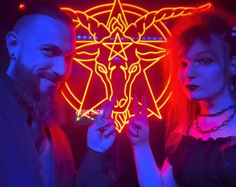 May include: Two people pose in front of a neon sign depicting a goat's head inside a pentagram. The sign is red and the people are illuminated in blue and red light. The sign reads "NEON SIGN TEXAS".