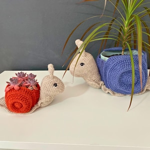 以下が含まれることがあります： 赤い貝殻と紫の貝殻の2つの編みぐるみカタツムリプランター。赤いカタツムリプランターには小さな多肉植物が入っています。紫のカタツムリプランターには緑の植物が入っています。