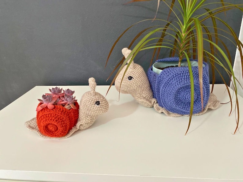 以下が含まれることがあります： 赤い貝殻と紫の貝殻の2つの編みぐるみカタツムリプランター。赤いカタツムリプランターには小さな多肉植物が入っています。紫のカタツムリプランターには緑の植物が入っています。