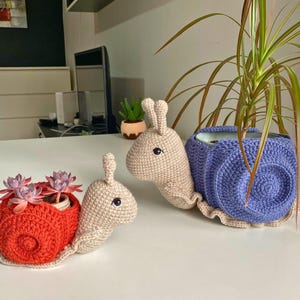 以下が含まれることがあります： 赤い殻と紫の殻の2つの編みぐるみカタツムリプランター。どちらもベージュの体と黒い目。赤いカタツムリプランターには小さな多肉植物が入っています。