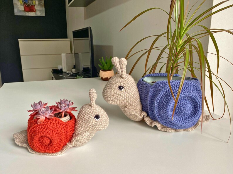以下が含まれることがあります： 赤い殻と紫の殻の2つの編みぐるみカタツムリプランター。どちらもベージュの体と黒い目。赤いカタツムリプランターには小さな多肉植物が入っています。
