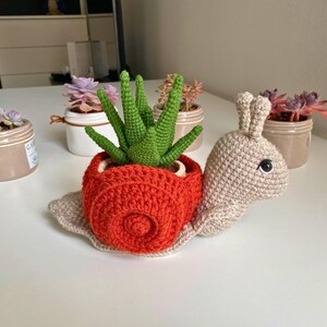 以下が含まれることがあります： 緑の多肉植物が入った、かぎ針編みで作ったカタツムリ型のプランター。カタツムリはベージュで、赤い殻と黒い目があります。