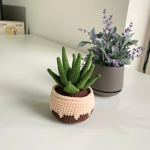 May include: A crocheted succulent plant in a brown and beige pot. The plant is green and has a spiky texture. The pot is sitting on a white table.
