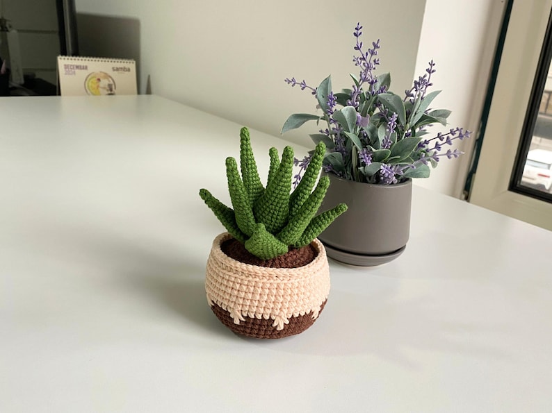 May include: A crocheted succulent plant in a brown and beige pot. The plant is green and has a spiky texture. The pot is sitting on a white table.