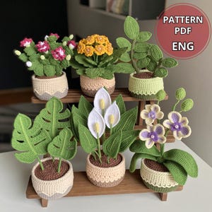 May include: Six crocheted potted plants arranged on a wooden tiered stand. The collection includes a pink flower plant, a yellow flower plant, a green leafy plant, a monstera, a white peace lily, and a purple and white orchid. Text reads PATTERN PDF ENG.