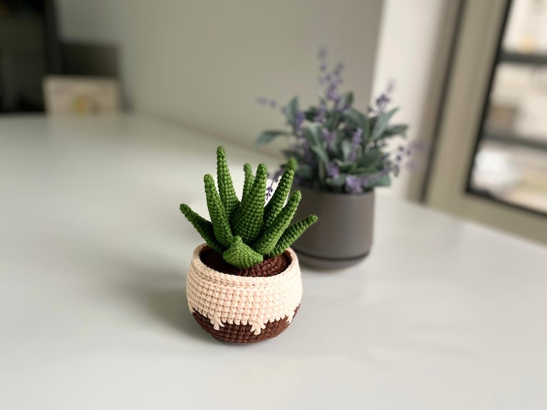 May include: A crocheted succulent plant in a brown and cream pot. The plant has green leaves and is sitting on a white surface.