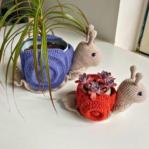 以下が含まれることがあります： 2つの編みぐるみカタツムリプランター。1つは薄い茶色のカタツムリで紫色のプランター、もう1つは薄い茶色のカタツムリで赤いプランター。両方のカタツムリは黒い目をしていて、小さな植物を持っています。