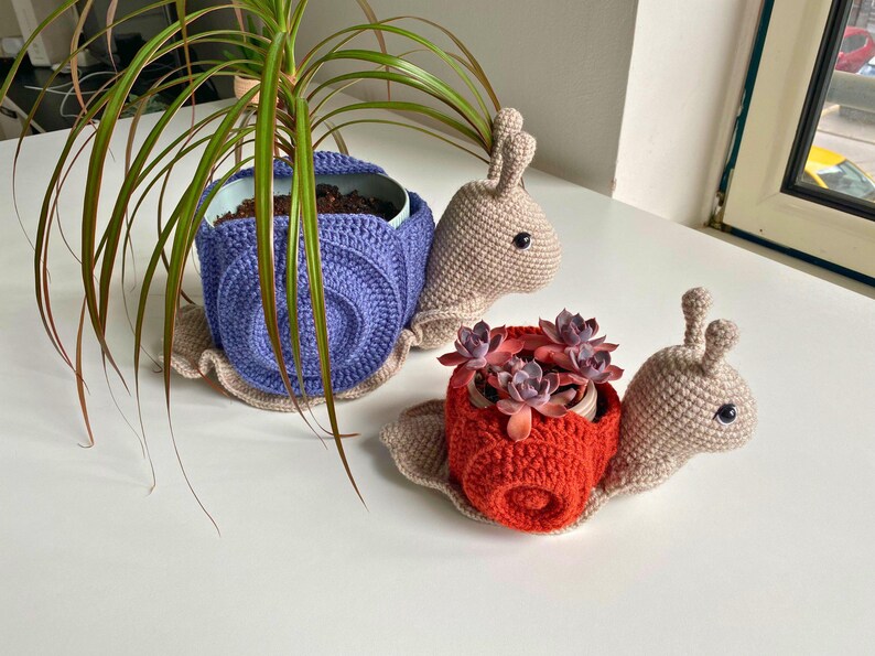以下が含まれることがあります： 2つの編みぐるみカタツムリプランター。1つは薄い茶色のカタツムリで紫色のプランター、もう1つは薄い茶色のカタツムリで赤いプランター。両方のカタツムリは黒い目をしていて、小さな植物を持っています。