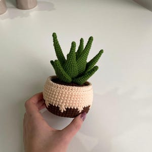 May include: A crocheted succulent plant in a brown and cream pot. The plant has green leaves and is sitting on a white surface.