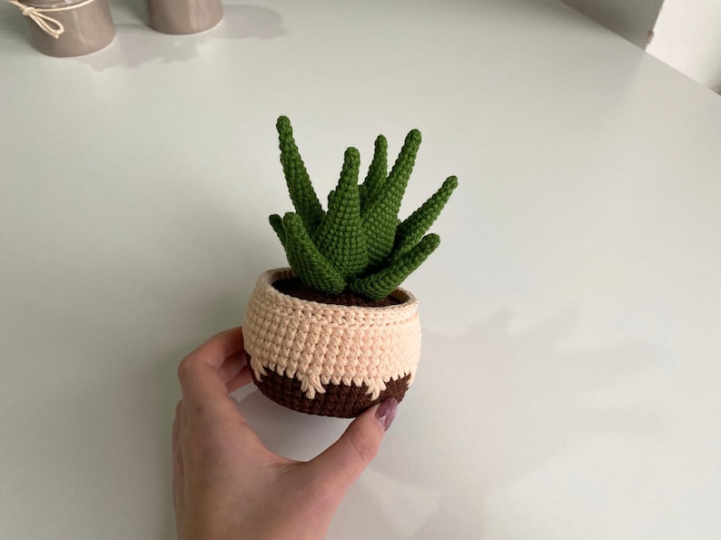 May include: A crocheted succulent plant in a brown and cream pot. The plant has green leaves and is sitting on a white surface.