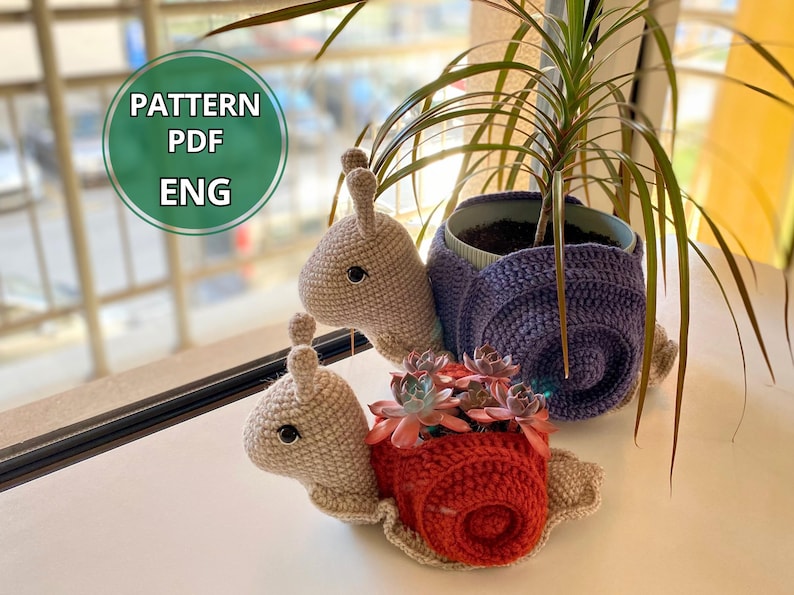 以下が含まれることがあります： 紫と赤の2つの編みぐるみカタツムリプランター。赤いカタツムリプランターには小さな多肉植物が入っています。カタツムリは白い表面に置かれ、背景には緑の植物があります。