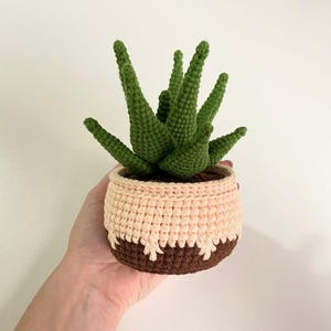May include: A crocheted succulent plant in a brown and pink pot. The plant has green leaves and is sitting on a white surface.