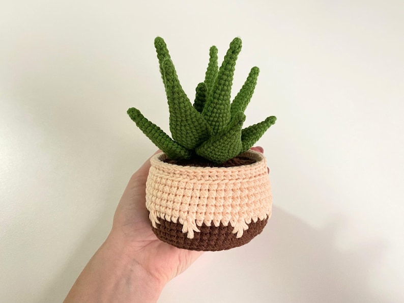May include: A crocheted succulent plant in a brown and pink pot. The plant has green leaves and is sitting on a white surface.