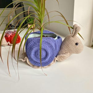 以下が含まれることがあります： 紫色の殻と赤い殻の2つの編みぐるみカタツムリプランター。両方のカタツムリはベージュの体で、小さな鉢植えの植物を持っています。