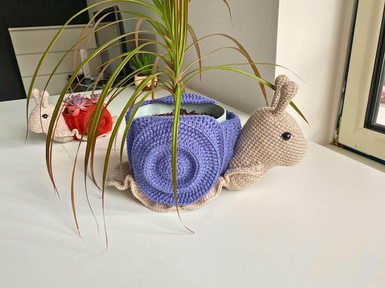 以下が含まれることがあります： 紫色の殻と赤い殻の2つの編みぐるみカタツムリプランター。両方のカタツムリはベージュの体で、小さな鉢植えの植物を持っています。