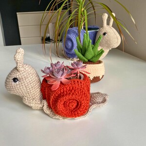 以下が含まれることがあります： 多肉植物が入った、かぎ針編みで作った3つのカタツムリ型のプランター。カタツムリはベージュ色で、茶色の目と赤、青、ベージュの殻をしています。