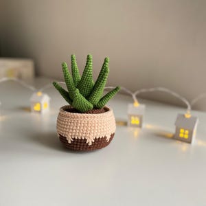 May include: A crocheted succulent plant in a brown and beige pot. The plant has green leaves and is sitting on a white surface with a string of white lights in the background.