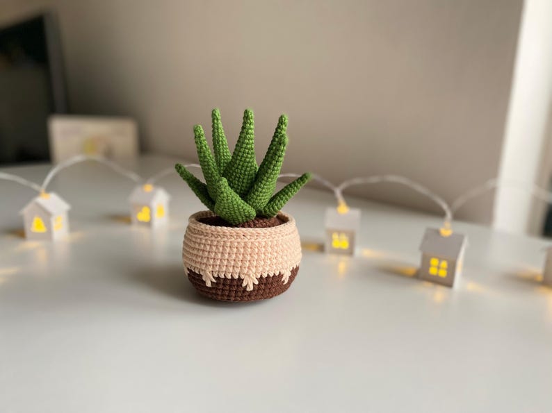 May include: A crocheted succulent plant in a brown and beige pot. The plant has green leaves and is sitting on a white surface with a string of white lights in the background.
