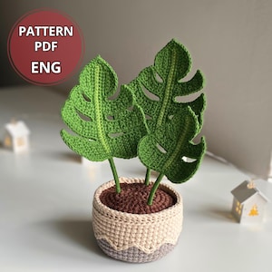 May include: A crocheted Monstera plant with three large green leaves in a beige and brown pot. The pot has a decorative wavy trim. The text "PATTERN PDF ENG" is in a red circle in the upper left corner.