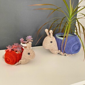 以下が含まれることがあります： 赤い鉢と紫の鉢の2つの編み物でできたカタツムリ型のプランター。中には小さな植物が入っています。カタツムリは白い表面の上に置かれています。