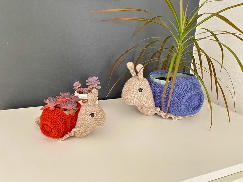 以下が含まれることがあります： 赤い鉢と紫の鉢の2つの編み物でできたカタツムリ型のプランター。中には小さな植物が入っています。カタツムリは白い表面の上に置かれています。