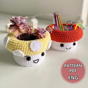 May include: Two crocheted mushroom-shaped planters, one yellow and white with a succulent plant inside, the other red and white with colorful pencils inside. Both planters have black eyes and a smiling mouth. The text "PATTERN PDF ENG" is on a pink circle.