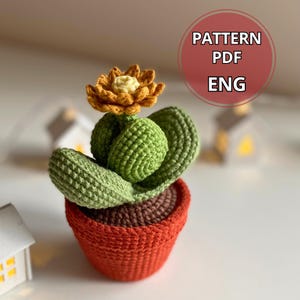 May include: A handmade crochet cactus in a terracotta-colored pot. The cactus is green with a yellow and orange flower. The pot is a reddish-orange color. The image includes the text "PATTERN PDF ENG" in a red circle.