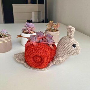 以下が含まれることがあります： 赤い殻とベージュの体をした、かぎ針編みで作ったカタツムリ型のプランター。カタツムリは、多肉植物が入った小さな鉢を持っています。