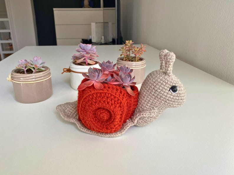 以下が含まれることがあります： 赤い殻とベージュの体をした、かぎ針編みで作ったカタツムリ型のプランター。カタツムリは、多肉植物が入った小さな鉢を持っています。