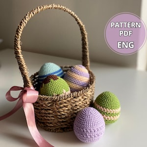 Pode incluir: Uma cesta de vime com uma alça de fita rosa cheia de ovos de Páscoa de crochê em várias cores, incluindo verde, roxo, azul e branco. A cesta está sobre uma superfície branca com o texto "PATTERN PDF ENG" em um círculo rosa.