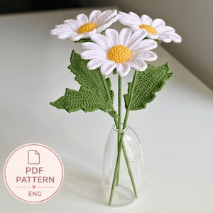 Puede incluir: Tres flores de margarita tejidas a ganchillo con pétalos blancos y centros amarillos, cada una unida a un tallo verde con hojas verdes dentadas. Las flores están dispuestas en un florero de vidrio transparente. Una insignia muestra PDF PATTERN ENG.