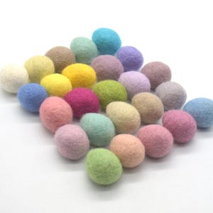 Puede incluir: Una colección de pequeñas bolas de fieltro redondas en una variedad de colores pastel y vibrantes, incluyendo rosa, amarillo, azul, verde, morado y beige. Las esferas están dispuestas en un grupo suelto y ligeramente superpuesto sobre una superficie blanca.