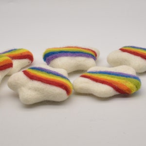 Może przedstawiać: Kilka białych, filcowych ozdób w kształcie chmur z tęczowym wzorem. Tęczowe paski są czerwone, pomarańczowe, żółte, zielone, niebieskie i fioletowe. Ozdoby ułożone są na białym tle.