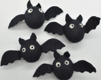 May include: Four small, black, felted wool bat figures arranged in a group. Each bat features a rounded body, two pointed ears, and two wide, white circular eyes with black pupils. The wings are extended outward with scalloped edges.