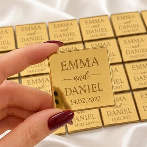 May include: A square gold-colored acrylic plaque with a reflective surface. The text EMMA and DANIEL is engraved in the center, followed by the date 14.02.2027. Multiple identical plaques are arranged in a grid pattern on a white fabric surface.