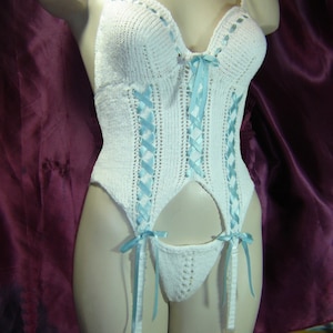 Op de afbeelding: Witte gehaakte lingerie set met lichtblauwe lintaccenten. De set bevat een bustier, jarretelgordel en string. De bustier heeft een sweetheart halslijn en lintvetersluiting. De jarretelgordel heeft bevestigde jarretels. De lingerie wordt getoond op een mannequin.