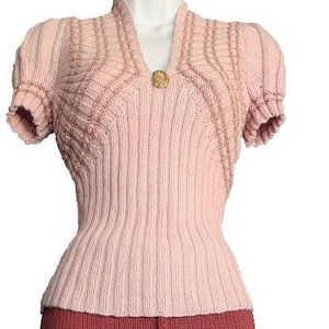 Puede incluir: Un suéter de punto rosa con cuello en V y mangas cortas. El suéter tiene un patrón decorativo y un botón dorado en el cuello.