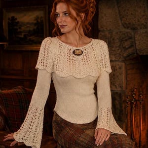 May include: Cream-coloured knitted sweater with a lace-like cape detail and long sleeves with flared cuffs. A decorative brooch is pinned at the neckline. The model is wearing a plaid skirt.