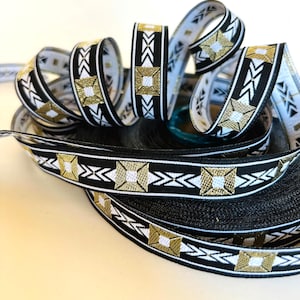 May include: Decorative ribbon featuring a repeating geometric pattern in black, white, and gold. The ribbon is coiled and layered, displaying the intricate design. The pattern includes squares and arrow-like shapes, creating a visually appealing texture. Ideal for crafts and gift wrapping.