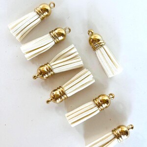 May include: Close-up of several cream-colored tassels with gold-colored metal caps and loops. The tassels are made of a soft, fringed material. The gold caps have a rounded shape.