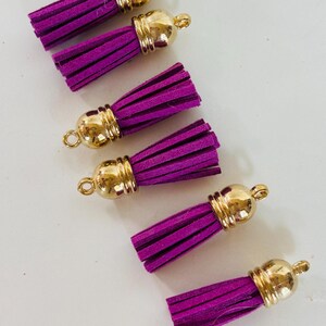 May include: Six purple suede tassels with gold-colored metal caps. The tassels are arranged on a white surface. Each tassel has a loop at the top for easy attachment to jewelry or other crafts.