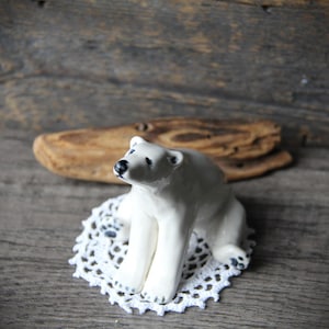Peut inclure: Figurine d'ours polaire en céramique blanche avec des détails noirs sur le nez et les pattes. L'ours est posé sur un napperon blanc crocheté. Le fond présente un morceau de bois flotté sur une surface en bois.