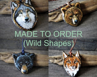 May include: Four ceramic animal head pendants on a wooden background. The pendants are of a wolf, a bear, a black bear, and a lynx. Each pendant is on a cord with a loop at the top. The text "MADE TO ORDER (Wild Shapes)" is printed below the pendants.