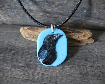 May include: A black raven pendant necklace featuring a detailed bird portrait on a light blue oval background. The pendant hangs from a black braided cord. The necklace is displayed on a wooden surface.