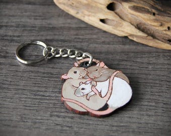 Könnte beinhalten: Ein Holzschlüsselanhänger mit einem gemalten Bild von drei Ratten, die sich aneinanderkuscheln. Die Ratten sind braun, grau und weiß. Der Schlüsselanhänger ist an einem silbernen Metallring befestigt.