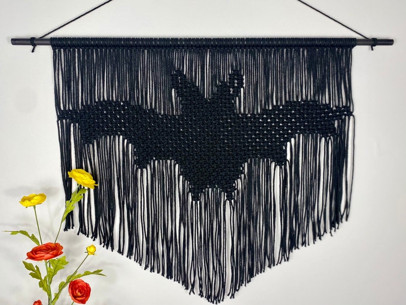 May include: Black macrame wall hanging featuring a bat design. The hanging has a fringe border and is made with black yarn.